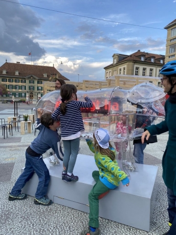 A giant transparent Pig is standing in the middle of a big open public square surrounded by buildings. A group of three children and one adult surround Pig, looking at it and touching it.
